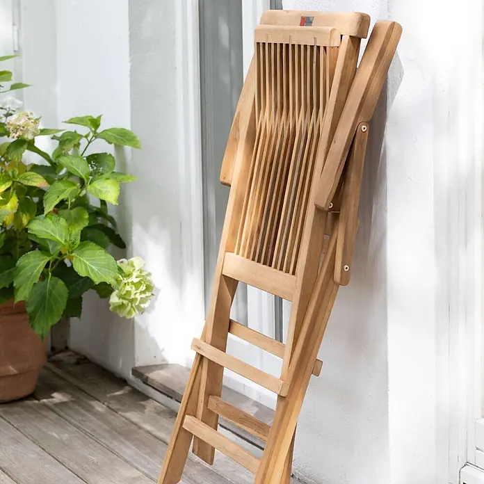Bene Living Gartenstuhl Andover - 1 Stk., Teakholz, Klappbar, Natur - Ein zusammengeklappter Gartenstuhl aus Teakholz lehnt an einer weißen Wand auf einer Holzterrasse neben einer Topfpflanze.