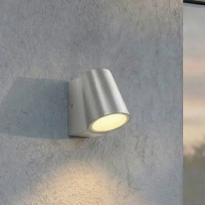 Lindby Außenwandleuchte Illuvia - alu / grau / zink, alu / grau / zink, IP44 - Gebürstete Metall-Wandleuchte, die warmes Licht nach unten abstrahlt, an einer strukturierten grauen Wand montiert.