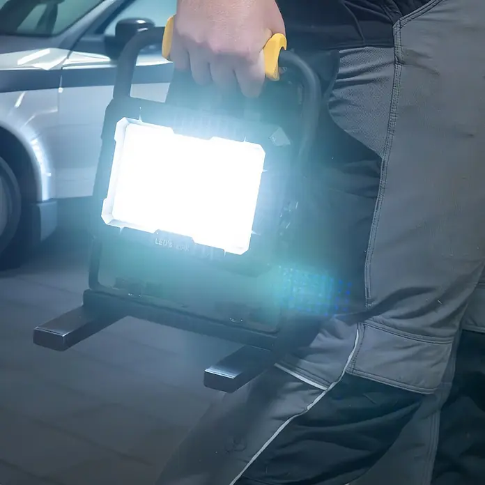 LED's work LED-Strahler für 18 V Akkus - 2.000 lm, 5.000 K, L x B x H: 214 x 220 x 309 mm, IP20 - Eine Person in Arbeitshosen hält eine schwarze tragbare LED-Arbeitsleuchte mit gelbem Griff, die den Bereich hell beleuchtet.