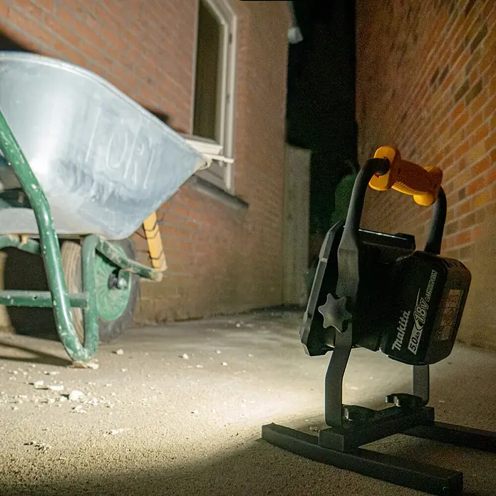 LED's work LED-Strahler für 18 V Akkus - 2.000 lm, 5.000 K, L x B x H: 214 x 220 x 309 mm, IP20 - Makita 18V LED Arbeitsleuchte beleuchtet einen Bereich neben einer Schubkarre und Ziegelwand.