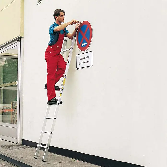 Zarges Anlegeleiter Anlegeleiter Alto L 10 Sprossen - Anzahl Tritte: 10 Stk., Arbeitshöhe: 3.900 m, Aluminium - Silberne Aluminiumleiter, von einem Mann zum Anbringen eines Schildes an der Wand verwendet.