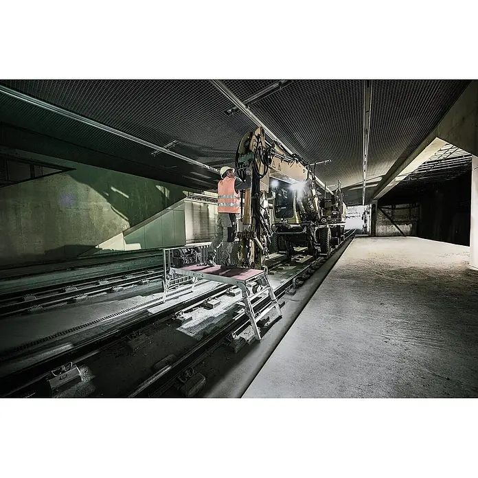 Munk Arbeitsplattform ML Maschinenpodest höhenverstellbar - Anzahl Tritte: 3 Stk., Arbeitshöhe: 2.730 m, Aluminium - Arbeiter auf einer silbernen, klappbaren Arbeitsplattform mit brauner Oberseite, der Wartungsarbeiten an einer gelben Baumaschine auf Gleisen durchführt.