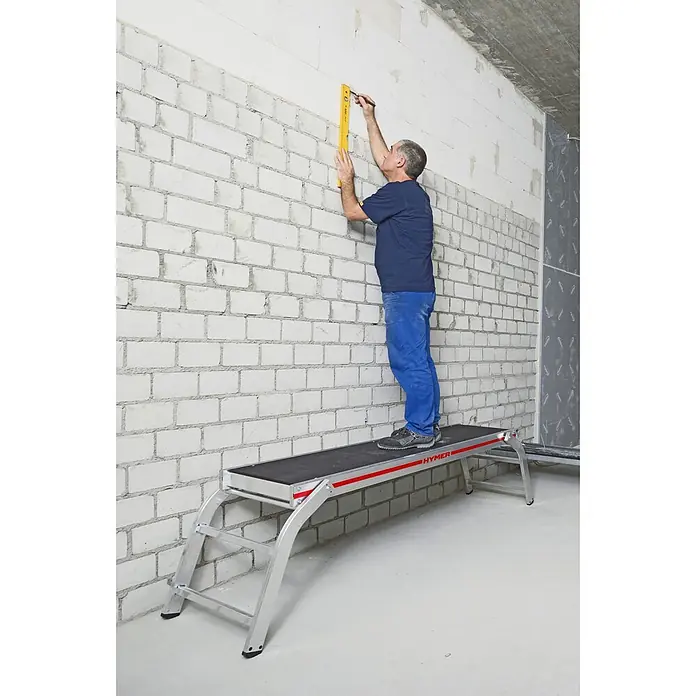 Hymer Arbeitsplattform Montageplattform Arbeitsplattform mit verschraubten Leiternfüßen - Anzahl Tritte: 2 Stk., Arbeitshöhe: 2.580 m, Aluminium - Mann steht auf einer silbernen Alu-Arbeitsplattform mit schwarzer Antirutschfläche und rotem HYMER-Streifen, markiert eine weiße Ziegelwand.