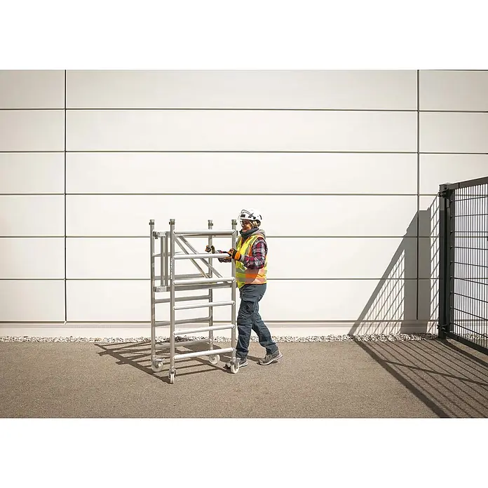 Werner Rollgerüst Klappgerüst Tower 3T nach DIN Norm EN 1004-1 AH 7,55m - Arbeitshöhe: 7.550 m, Belastbarkeit Plattform: 216 kg - Eine Person in Schutzkleidung bewegt ein silbernes Aluminium-Rollgerüst, was seine Tragbarkeit hervorhebt.