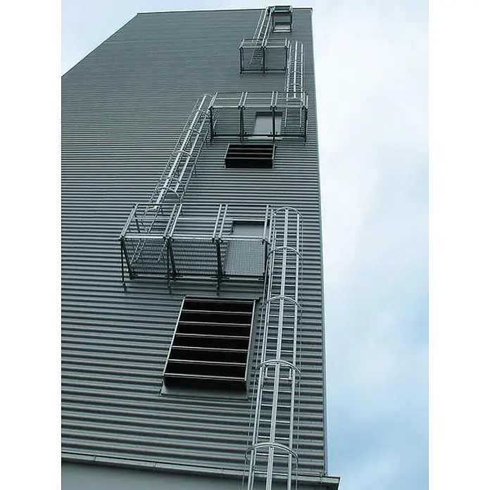 Munk Anlegeleiter ortsfeste Steigleitern Aluminium, 15,96m SH - Aluminium - Industrielle Steigleitern aus silbernem Metall mit Schutzkäfigen und Plattformen an einem Wellblechgebäude.