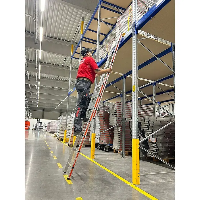Hymer Schiebeleiter Einhänge-Schiebeleiter mit Einhängehaken 2x8 Sprossen - Arbeitshöhe: 4.890 m, Aluminium - Person erklimmt eine Aluminiumleiter mit orangefarbenen Akzenten, um hohe Regale in einem Lager zu erreichen.