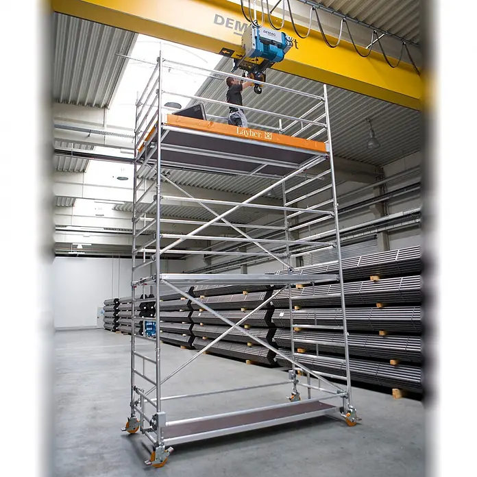 Layher Rollgerüst Rollgerüst Uni Breit P2 6,20m AH - Arbeitshöhe: 6.200 m - Hohes fahrbares Aluminiumgerüst mit dunklen Plattformen und orangefarbenen Rollen, ein Arbeiter auf der obersten Plattform wartet einen Kran.