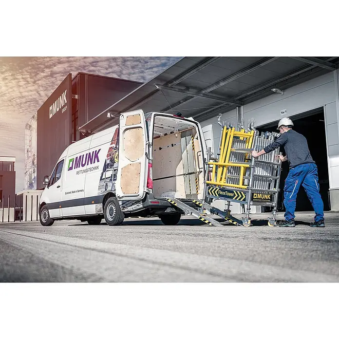 Munk Rollgerüst Flexxtower SG mit Sicherheitsgeländer 4,10m AH - Arbeitshöhe: 4.100 m - Ein Arbeiter lädt ein gelbes und silbernes FlexxTower Rollgerüst auf einen weißen Munk-Transporter über eine Rampe.
