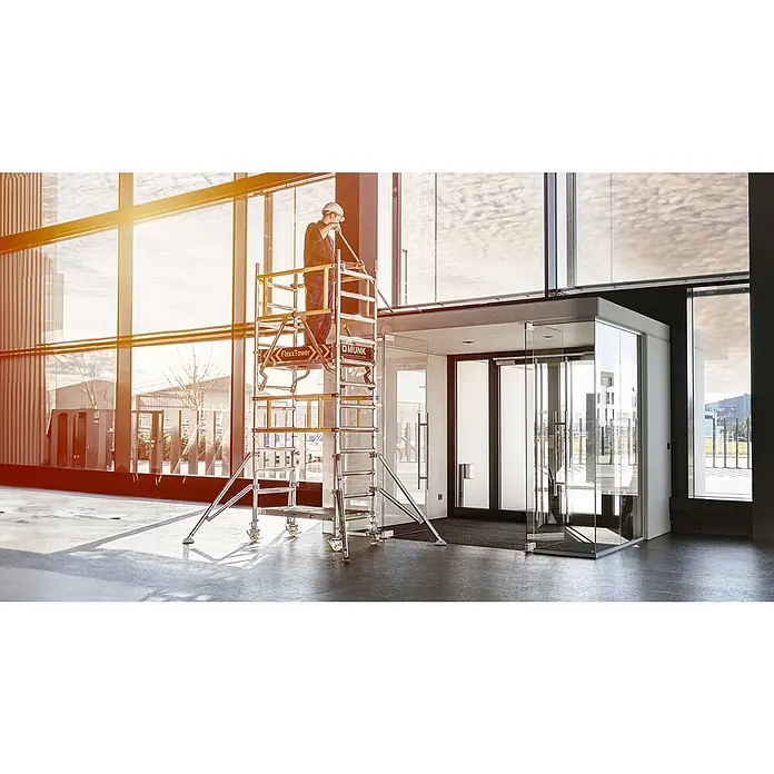 Munk Rollgerüst Flexxtower SG mit Sicherheitsgeländer 4,10m AH - Arbeitshöhe: 4.100 m - Arbeiter auf einem silbernen FlexxTower Rollgerüst mit gelben Akzenten, arbeitet an hohen Fenstern in einem modernen Gebäude.