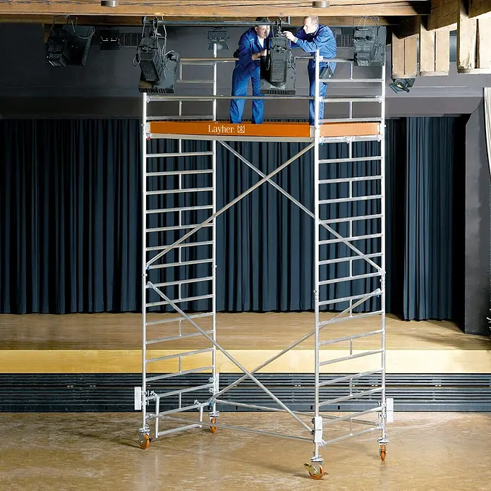 Layher Rollgerüst Rollgerüst Uni Breit 7,20m AH - Arbeitshöhe: 7.200 m - Aluminium-Rollgerüst mit orangefarbener Plattform, von zwei Arbeitern zum Einstellen von Bühnenscheinwerfern genutzt.