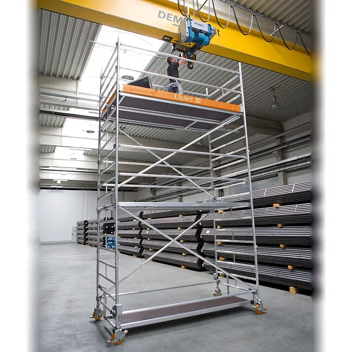 Layher Rollgerüst Rollgerüst Uni Breit 7,20m AH - Arbeitshöhe: 7.200 m - Ein mobiles Baugerüst aus silbernem Aluminium mit einer orangefarbenen Arbeitsplattform. Ein Arbeiter benutzt es an einem Kran in einer Industriehalle.