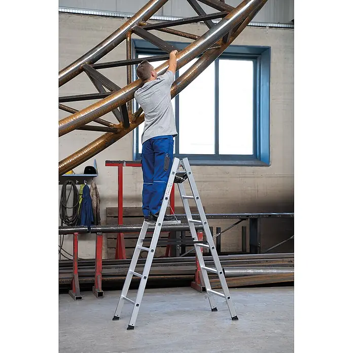 Munk Doppel-Stehleiter Sprossenstehleiter 2x10 Sprossen - Anzahl Tritte: 10 Stk., Arbeitshöhe: 4.200 m, Aluminium - Silberne Stehleiter, die ein Arbeiter benutzt, um eine Metallfachwerkkonstruktion in einer Werkstatt zu erreichen.
