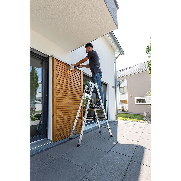 Munk Doppel-Stehleiter Sprossenstehleiter 2x10 Sprossen - Anzahl Tritte: 10 Stk., Arbeitshöhe: 4.200 m, Aluminium - Silberne Aluminium-Klappleiter, auf der ein Mann ein Außenlicht an einem modernen Haus befestigt.