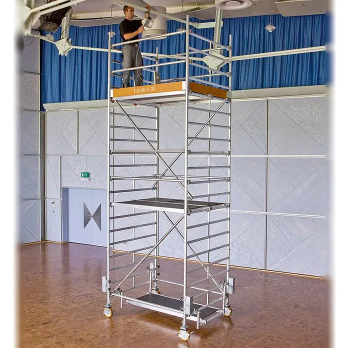 Layher Rollgerüst Rollgerüst Uni Kompakt 8,38m AH (DIN EN 1004:2004) - Arbeitshöhe: 8.380 m - Hohes, silbernes Rollgerüst mit einem Mann, der an einer Deckenhalterung arbeitet, zeigt dessen Höhe und Anwendung.