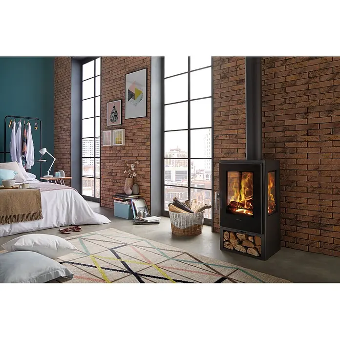 Panadero Kaminofen Andes 3V Ecodesign - 8 kW, Raumheizvermögen: 240 m³, Material Verkleidung: Stahl, Schwarz Metallic - Moderner schwarzer Holzkamin mit Glastür und Holzfach, brennt aktiv in einem gemütlichen Schlafzimmer mit Ziegelwänden und großen Fenstern.