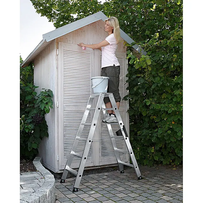 Munk Stehleiter Stehleiter mit ergo-pad beidseitig begehbar 2x3 Stufen - Anzahl Tritte: 3 Stk., Arbeitshöhe: 0,67 m, Belastbarkeit: 150 kg, Aluminium - Silberne Aluminiumleiter, von einer Frau zum Reinigen eines Gartenhauses verwendet, mit Eimer auf der Plattform.