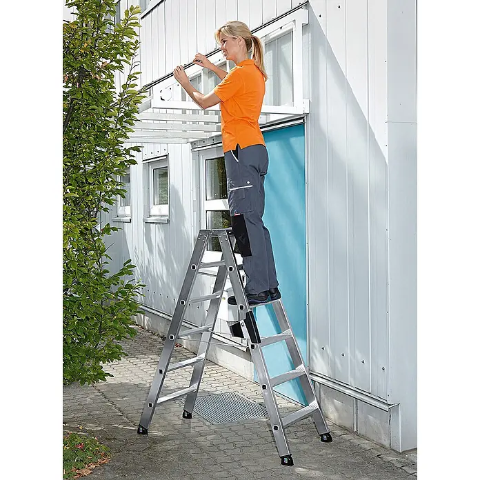 Munk Stehleiter Stehleiter beidseitig begehbar mit relax-step 2x14 Stufen - Anzahl Tritte: 6 Stk., Arbeitshöhe: 2.900 m, Aluminium - Frau auf einer silbernen Aluminiumleiter installiert ein Element über einer Gebäudetür.