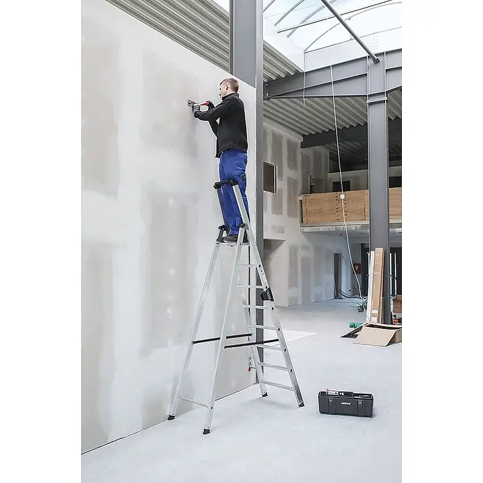 Munk Stehleiter Stehleiter mit clip-step 5 Stufen - Anzahl Tritte: 5 Stk., Arbeitshöhe: 3.150 m, Aluminium - Mann auf einer silbernen Aluminium-Klappleiter, der an einer Wand in einem Rohbau arbeitet.