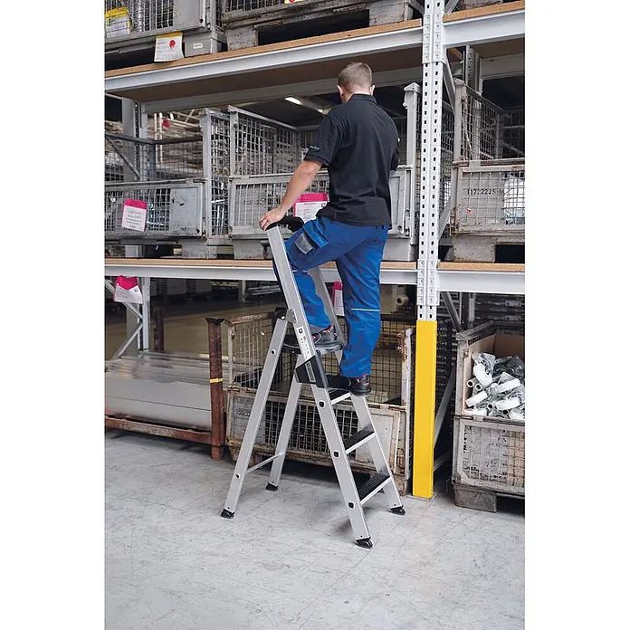 Munk Stehleiter Stehleiter mit clip-step 5 Stufen - Anzahl Tritte: 5 Stk., Arbeitshöhe: 3.150 m, Aluminium - Silberne Stehleiter mit schwarzen Stufen, von einer Person im Lager zum Erreichen von Regalen genutzt.