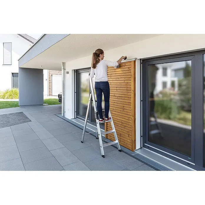 Munk Stehleiter Stehleiter mit clip-step 5 Stufen - Anzahl Tritte: 5 Stk., Arbeitshöhe: 3.150 m, Aluminium - Frau steht auf einer silbernen Aluminium-Stehleiter und arbeitet an einem hölzernen Sichtschutz.