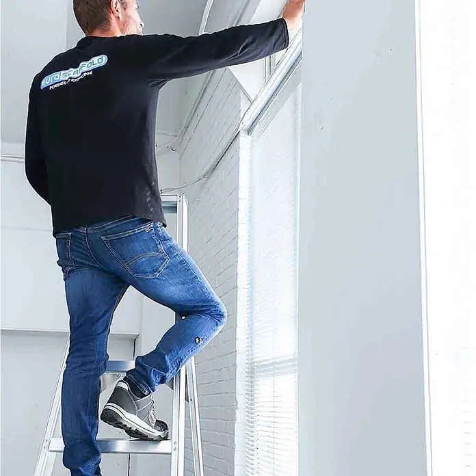 ASC Stehleiter Eurostairs Stufenstehleiter einseitig eloxiert mit Plattform 4 Stufen - Anzahl Tritte: 4 Stk., Arbeitshöhe: 2.800 m, Aluminium - Aluminium-Stehleiter, die eine Person beim Einstellen von Fensterjalousien stützt.