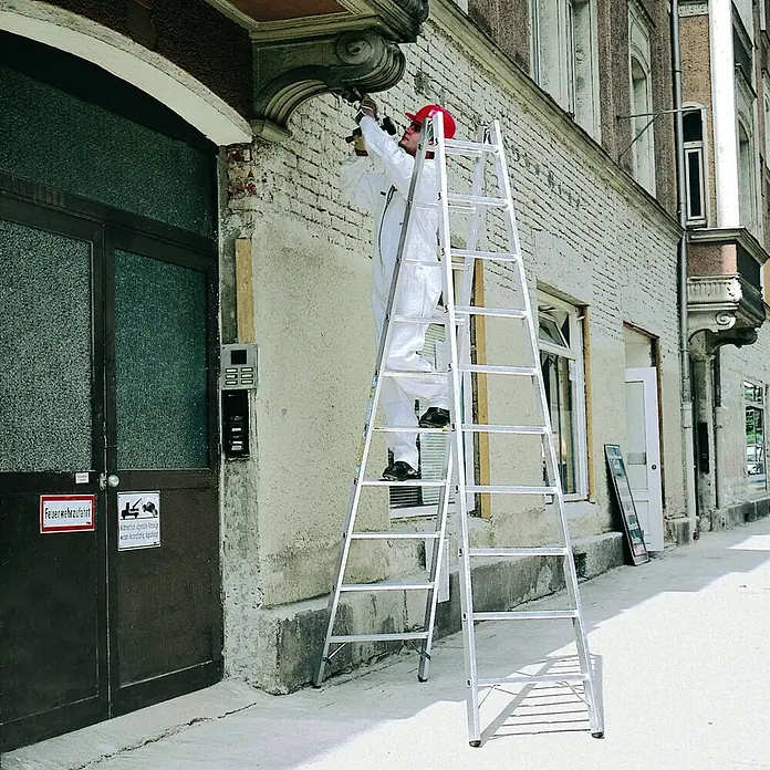 Zarges Stehleiter Sprossenstehleiter Coni B 2x18 Sprossen - Anzahl Tritte: 18 Stk., Arbeitshöhe: 6.150 m, Aluminium - Silberfarbene, ausziehbare Aluminiumleiter, von einem Arbeiter zur Reparatur einer alten Gebäudefassade eingesetzt.