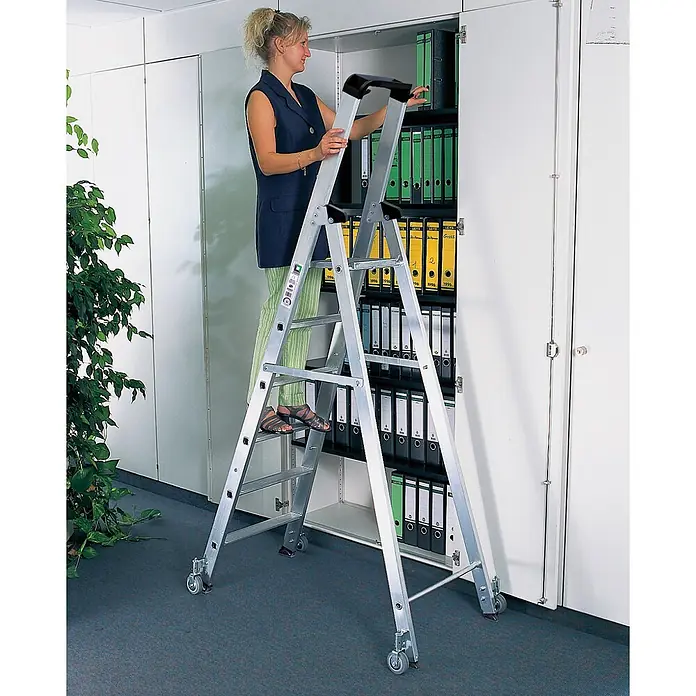 Munk Stehleiter Stehleiter einseitig begehbar mit Rollen 3 Stufen - Anzahl Tritte: 3 Stk., Arbeitshöhe: 2.700 m, Aluminium - Silberne Aluminium-Stehleiter mit Rollen. Eine Frau greift damit nach Akten in einem hohen Büroschrank.