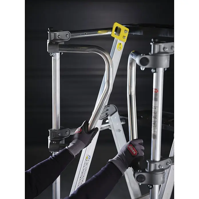 Altrex Podestleiter RolGuard Teleskop Plattformleiter Compact 5/7 Stufen - Arbeitshöhe: 3.600 m - Hände in Handschuhen montieren silberne Rohrschutzgeländer an ein modulares Leitersystem mit schwarzen Klemmen und gelbem Verschluss.
