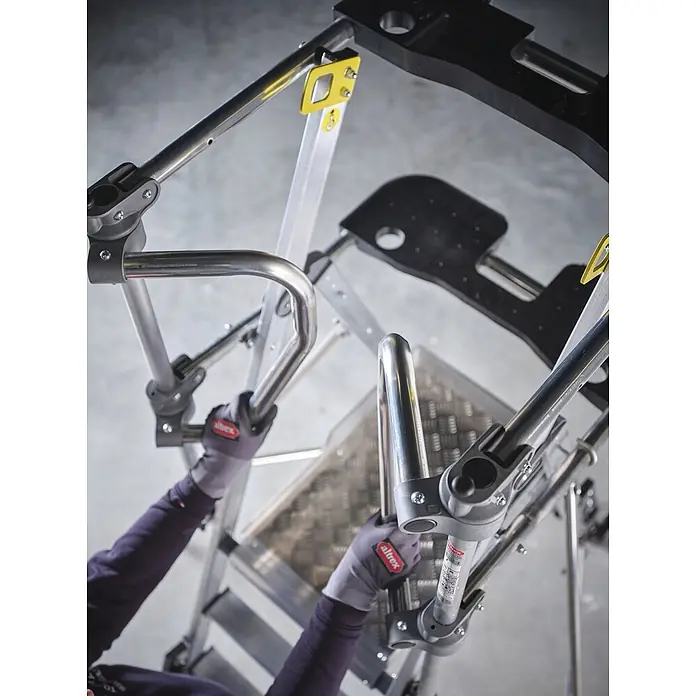 Altrex Podestleiter RolGuard Teleskop Plattformleiter Compact 5/7 Stufen - Arbeitshöhe: 3.600 m - Arbeiter steigt auf eine silberne Aluminium-Leiteranlage mit schwarzen Stufen und gelben Clips, hält sich an Metallhandläufen fest.