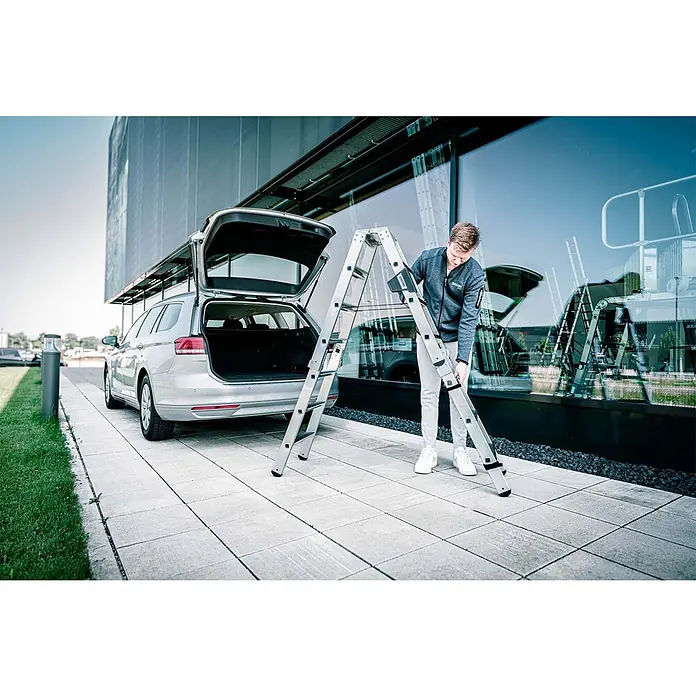Munk Stehleiter Stufen Stehleiter Set beidseitig begehbar steckbar inkl. Einsteckteil 2x7 S - Anzahl Tritte: 7 Stk., Arbeitshöhe: 3.150 m, Belastbarkeit: 150 kg - Mann faltet oder entfaltet eine silberne Teleskopleiter neben einem Auto.
