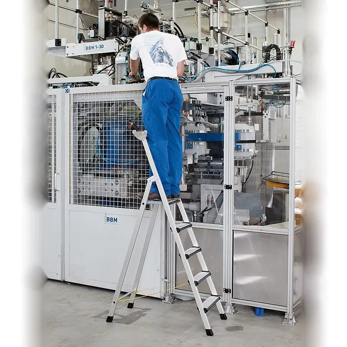 Zarges Stehleiter Comfortstep S gebördelte Stehleiter mit gepolsterter Stufen-Vorderkante 4 - Anzahl Tritte: 4 Stk., Arbeitshöhe: 2.900 m, Aluminium - Silberne Trittleiter mit schwarzen Stufen, wird von einem Mann an einer Industriemaschine benutzt.