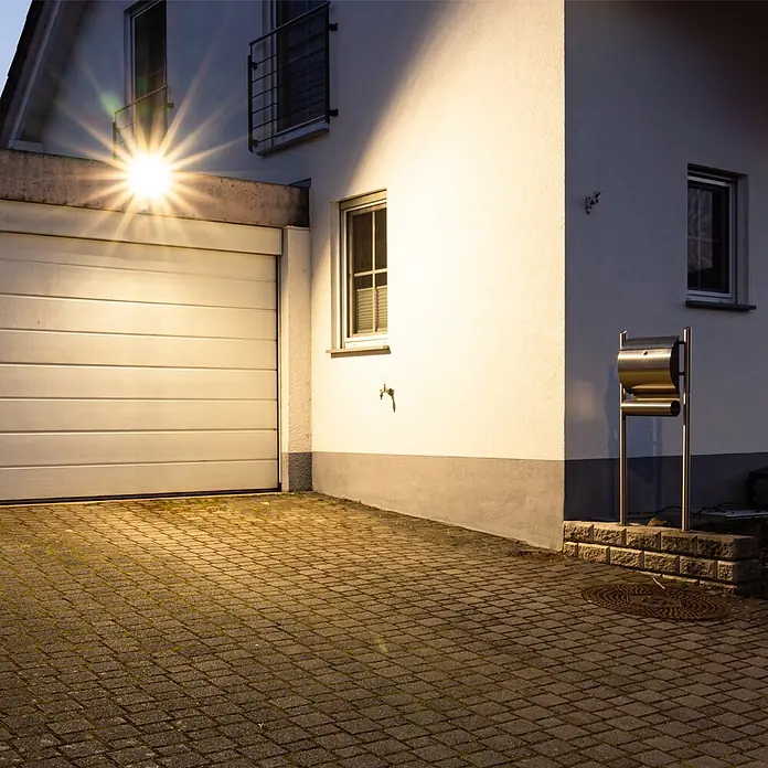 LED's work LED-Außenwandleuchte - 100 W, 4,7 x 24,1 x 26,6 cm, Schwarz, IP65 - Haus mit beleuchteter weißer Garage und gepflasterter Einfahrt bei Nacht, erhellt von einem hellen Außenlicht.