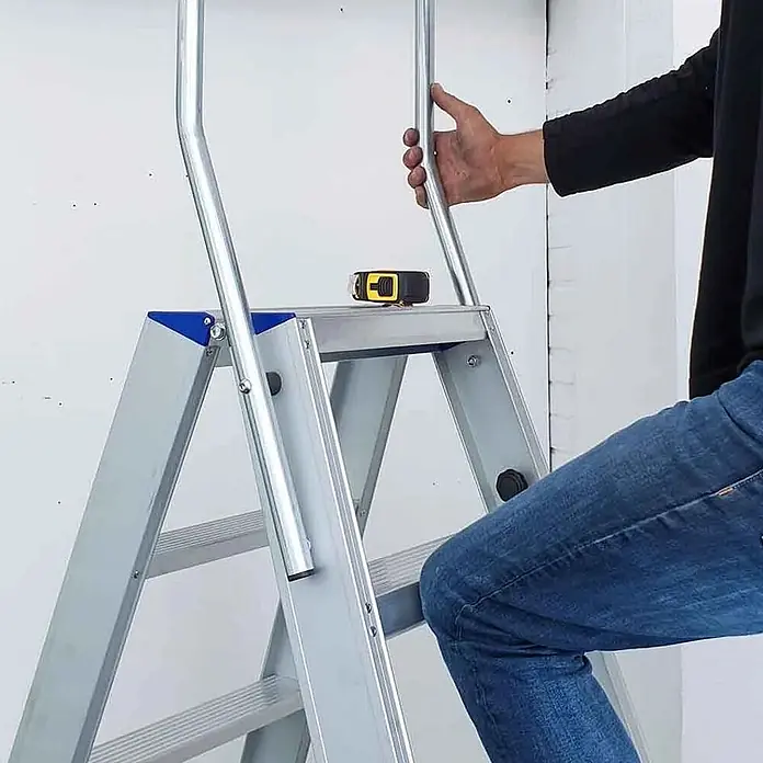 ASC Stehleiter Eurostairs Stufenstehleiter zweiseitig eloxiert mit Bügel 5 Stufen - Anzahl Tritte: 5 Stk., Arbeitshöhe: 3.050 m, Aluminium - Silberne Aluminium-Klappleiter mit blauen Akzenten, von einer Person am Handlauf benutzt. Ein Maßband liegt auf der obersten Stufe.