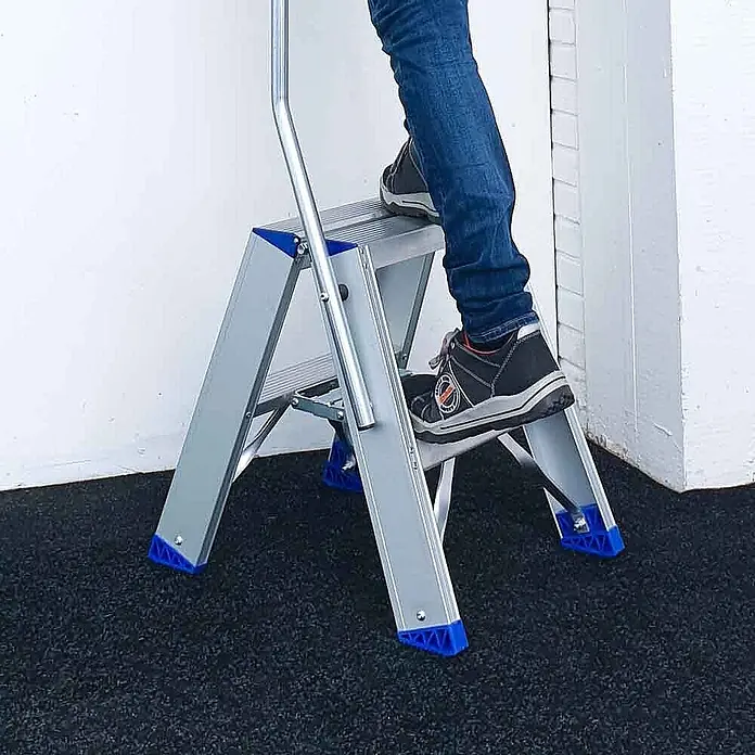 ASC Stehleiter Eurostairs Stufenstehleiter zweiseitig eloxiert mit Bügel 5 Stufen - Anzahl Tritte: 5 Stk., Arbeitshöhe: 3.050 m, Aluminium - Silberne Aluminium-Klappleiter mit blauen Akzenten. Eine Person steht auf den Stufen und demonstriert ihre Nutzung.