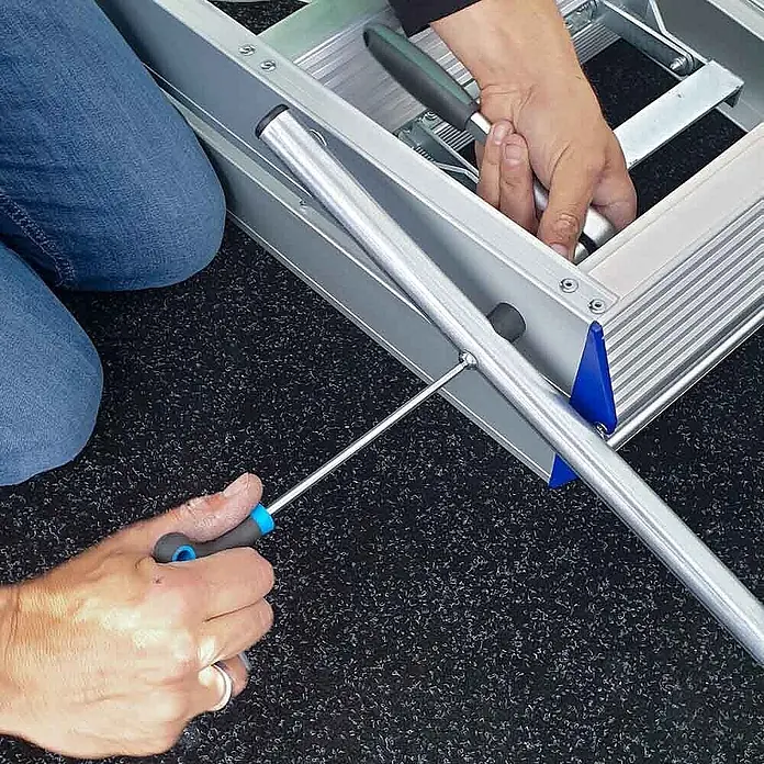 ASC Stehleiter Eurostairs Stufenstehleiter zweiseitig eloxiert mit Bügel 5 Stufen - Anzahl Tritte: 5 Stk., Arbeitshöhe: 3.050 m, Aluminium - Person montiert Aluminiumleiter mit Schraubendreher. Blaue Kunststoffteile sind am silbernen Rahmen sichtbar.