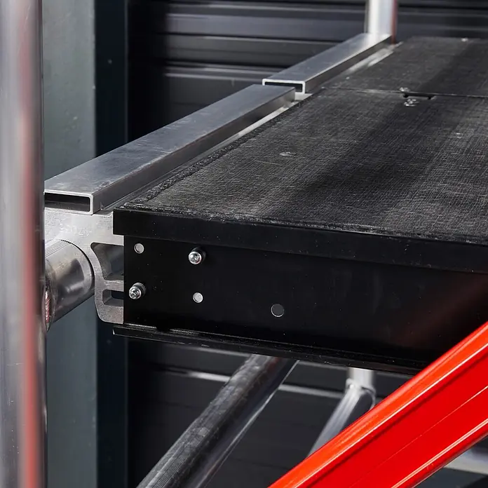 Altrex Rollgerüst RS Tower 55-S mit Safe-Quick® 6,8m Arbeitshöhe Holz-Plattform 2,45m - Arbeitshöhe: 6.800 m - Nahaufnahme einer schwarzen Gerüstplattform mit strukturierter Oberfläche, getragen von silbernen Aluminiumträgern und einer roten Metallstrebe.