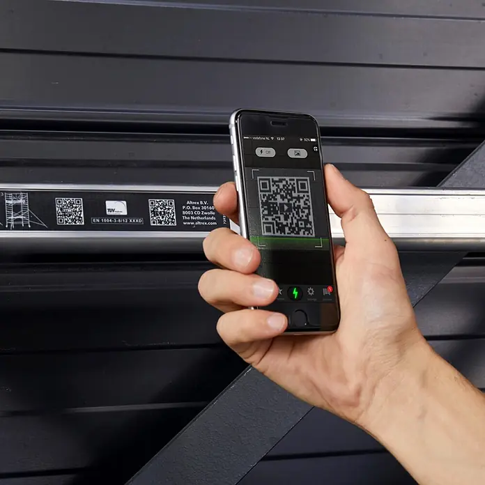 Altrex Rollgerüst RS Tower 55-S mit Safe-Quick® 6,8m Arbeitshöhe Holz-Plattform 2,45m - Arbeitshöhe: 6.800 m - Hand hält silbernes Smartphone, scannt QR-Code auf dunkler geriffelter Oberfläche.