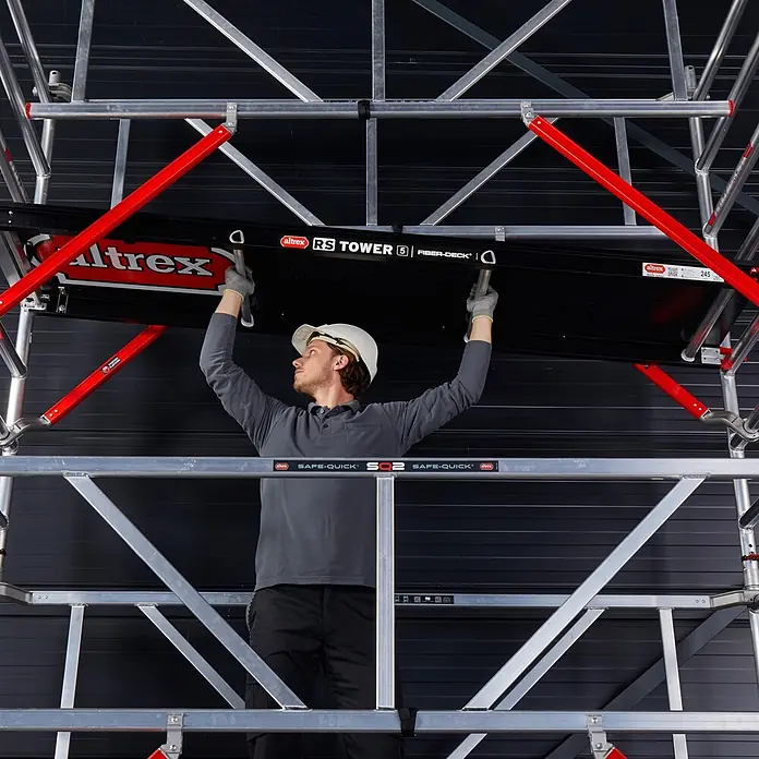 Altrex Rollgerüst RS Tower 55-S mit Safe-Quick® 6,8m Arbeitshöhe Holz-Plattform 2,45m - Arbeitshöhe: 6.800 m - Ein Mann montiert eine schwarze Fiber-Deck Plattform auf ein silbernes Altrex RS TOWER 5 Rollgerüst.