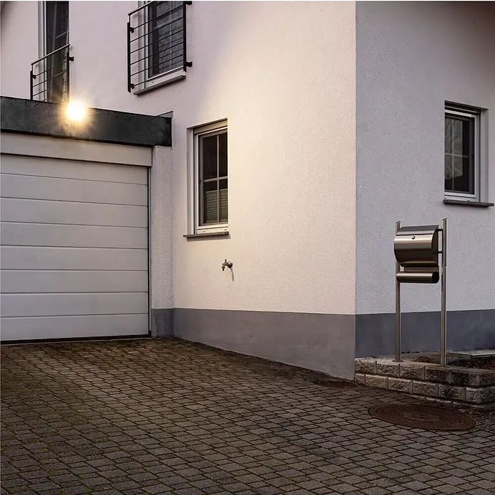 LED's work LED-Außenwandleuchte - 50 W, 3,7 x 17,9 x 19,9 cm, Schwarz, IP65 - Beleuchtete weiße Hausfassade mit geschlossener Garagentür, modernem Edelstahl-Briefkasten und dunkler gepflasterter Einfahrt.