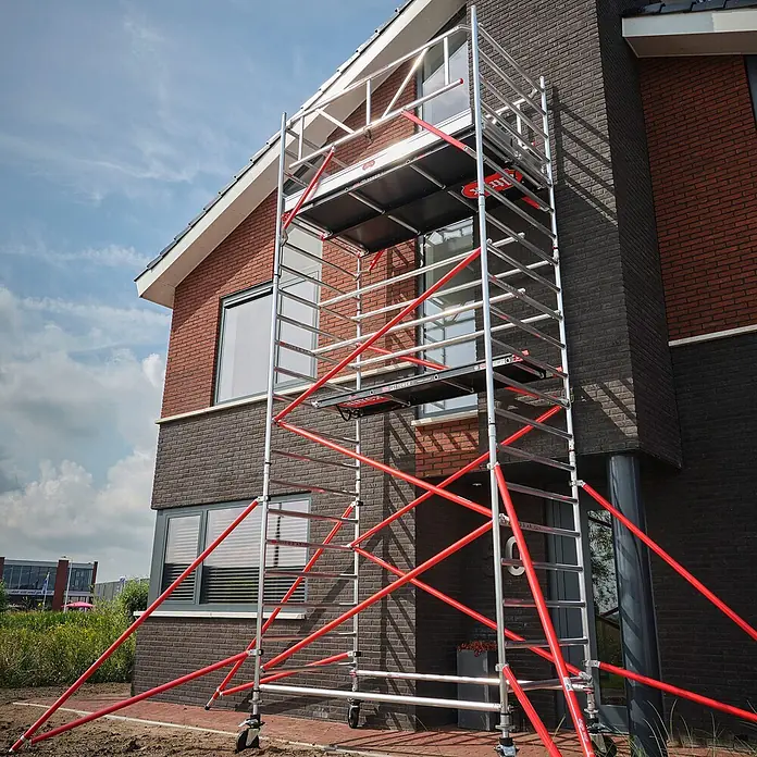 Altrex Rollgerüst Fahrgerüst RS Tower 52 Aluminium mit Fiber-Deck Plattform 13,20m AH 1,35x2 - Arbeitshöhe: 13.200 m - Mobiles Aluminiumgerüst mit roten Stützelementen, aufgebaut neben einem modernen Backsteinhaus.