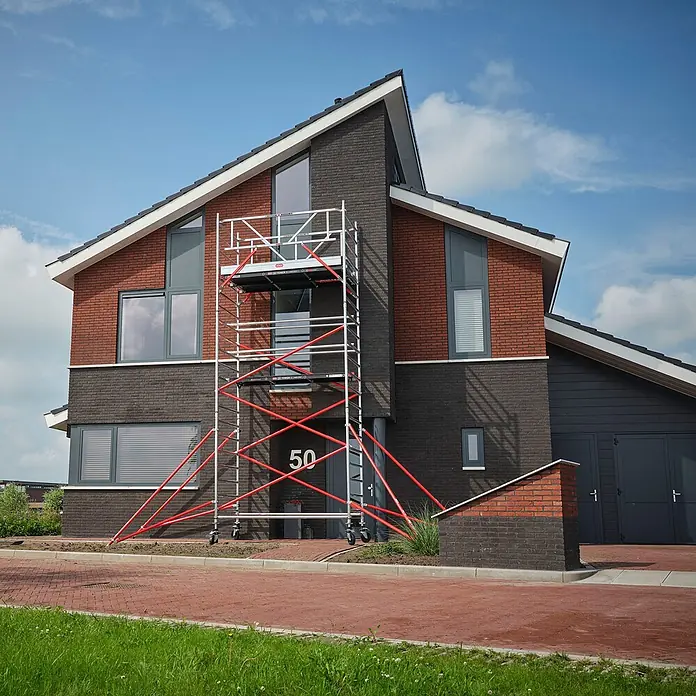 Altrex Rollgerüst Fahrgerüst RS Tower 52 Aluminium mit Fiber-Deck Plattform 13,20m AH 1,35x2 - Arbeitshöhe: 13.200 m - Silbernes Rollgerüst mit roten Diagonalstreben vor einem modernen Ziegelhaus.