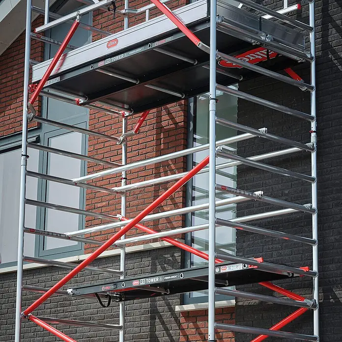 Altrex Rollgerüst Fahrgerüst RS Tower 52 Aluminium mit Fiber-Deck Plattform 13,20m AH 1,35x2 - Arbeitshöhe: 13.200 m - Mobiles Gerüst mit silbernem Metallrahmen, roten Diagonalstreben und schwarzen Plattformen vor einem Ziegelgebäude.