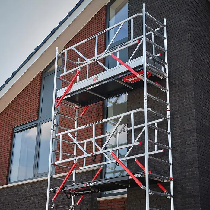 Altrex Rollgerüst Fahrgerüst RS Tower 51-S Safe-Quick Aluminium mit Fiber-Deck Plattform 10, - Arbeitshöhe: 10.200 m - Aluminiumgerüst mit roten Streben und Plattformen, an einem modernen Backsteinhaus für Arbeiten in der Höhe aufgestellt.