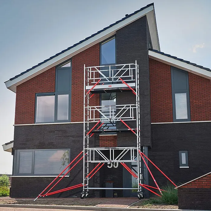 Altrex Rollgerüst Fahrgerüst RS Tower 51-S Safe-Quick Aluminium mit Fiber-Deck Plattform 10, - Arbeitshöhe: 10.200 m - Silberfarbenes Fahrgerüst mit roten Streben vor einem modernen Backsteinhaus.