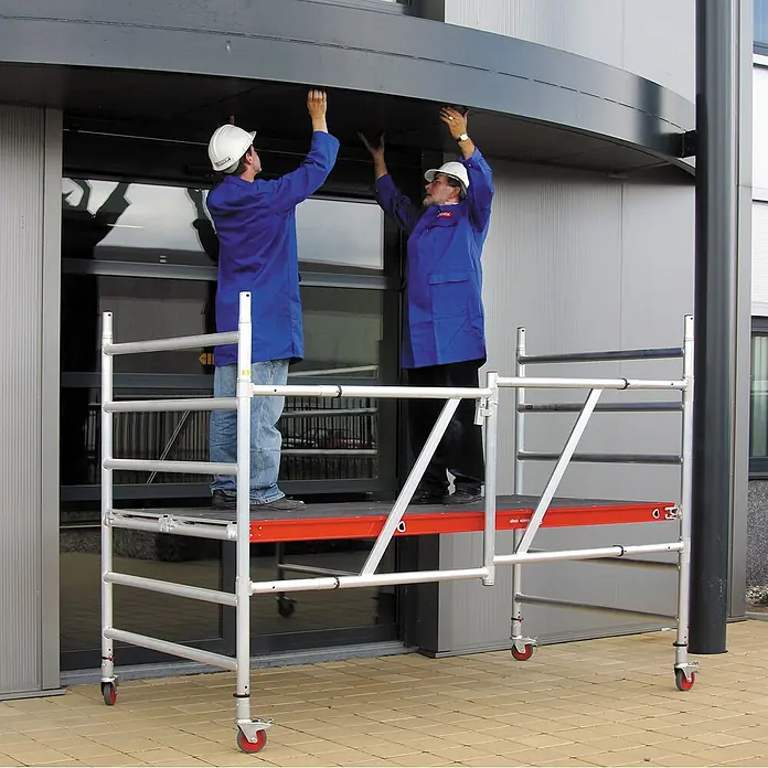 Altrex Rollgerüst Klappgerüst RS Tower 54 Alu Holz-Plattform 0,75x1,85m mit Safe-Quick 8,8m - Arbeitshöhe: 8.800 m - Zwei Arbeiter auf einem mobilen Aluminiumgerüst mit roten und schwarzen Plattformen, die eine Gebäudevorsprung erreichen.