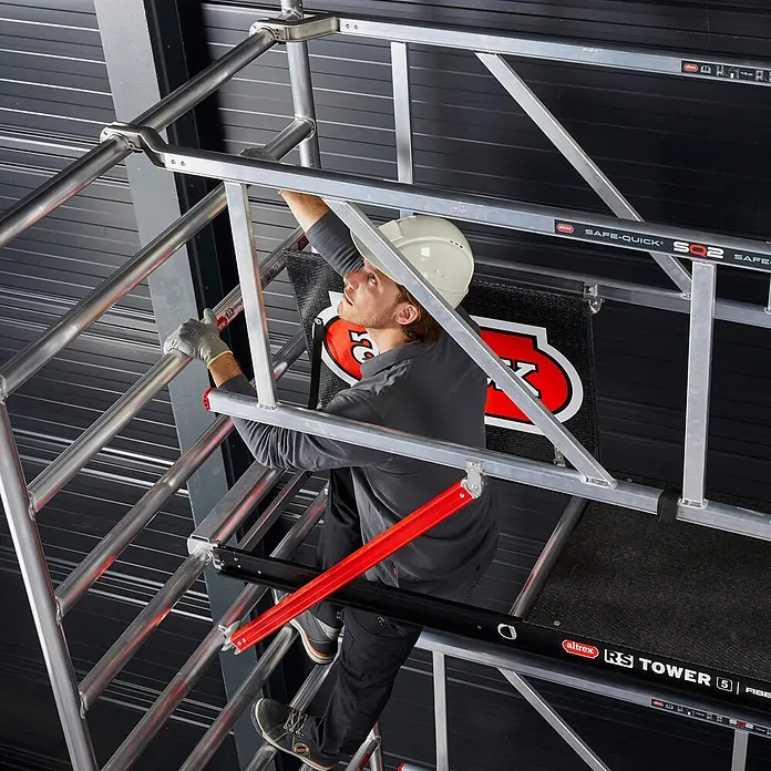 Altrex Rollgerüst Fahrgerüst RS Tower 52-S Aluminium mit Safe-Quick und Fiber-Deck Plattform - Arbeitshöhe: 12.200 m - Arbeiter klettert auf einen silbernen Aluminium Altrex Gerüstturm mit roten Akzenten.