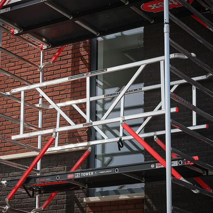Altrex Rollgerüst Fahrgerüst RS Tower 52-S Aluminium mit Safe-Quick und Fiber-Deck Plattform - Arbeitshöhe: 12.200 m - Altrex RS Tower 5 Rollgerüst mit silbernem Aluminiumrahmen, roten Streben und schwarzen Plattformen vor einer Ziegelwand.