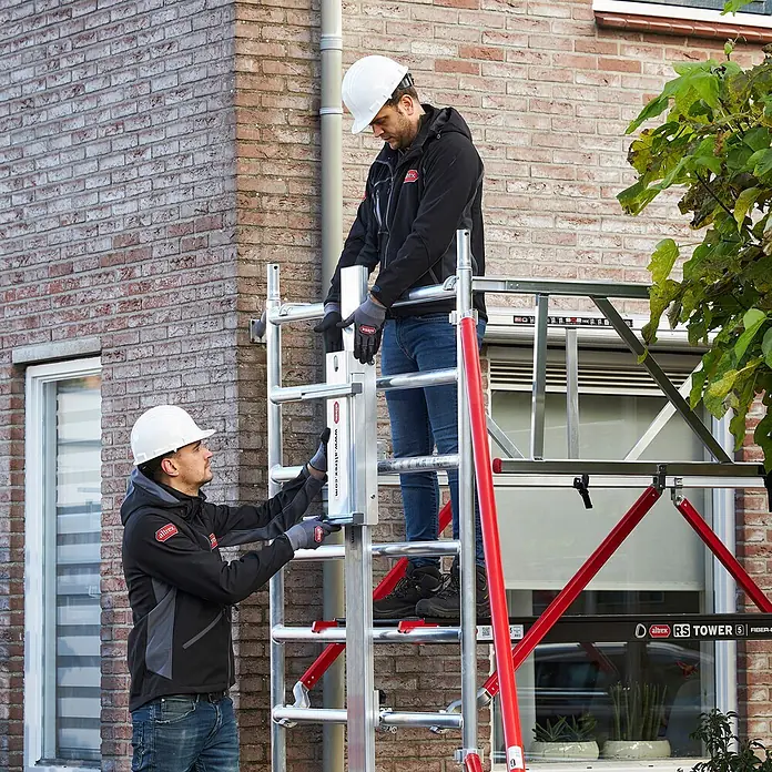 Altrex Rollgerüst Shuttle Basisset mit Winde 3,20m Plattformhöhe 5,20m Arbeitshöhe (kein Ge (Arbeitshöhe: 5.200 m, Belastbarkeit Plattform: 30 kg) -  - Zwei Arbeiter montieren ein silber-rotes Altrex RS Tower 5 Gerüst an einem Backsteinhaus.