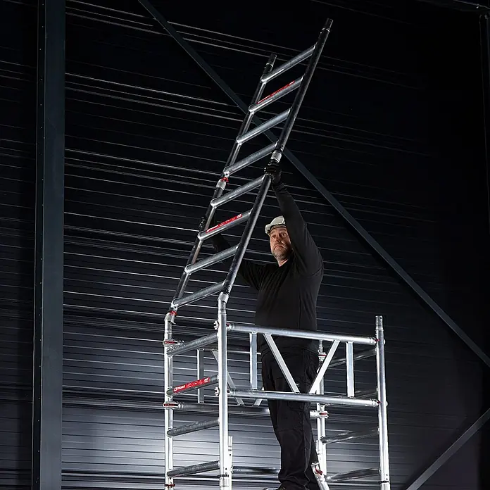Altrex Rollgerüst MiTower 1-Personen-Gerüst Aluminium mit Holz-Plattform 0,75x1,27m 5,20m AH - Arbeitshöhe: 5.200 m - Arbeiter hebt ein silbernes Aluminium-Leiterteil für ein mobiles Gerüst, zeigt die Montage.