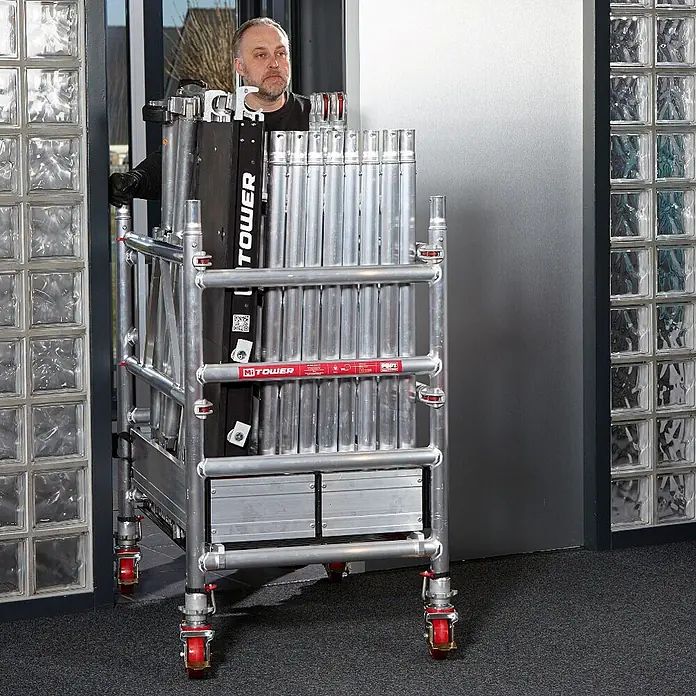 Altrex Rollgerüst MiTower 1-Personen-Gerüst Aluminium mit Holz-Plattform 0,75x1,27m 5,20m AH - Arbeitshöhe: 5.200 m - Kompaktes, silbernes Mİ TOWER Rollgerüst, von einem Mann auf roten Rädern im Innenbereich geschoben, transportbereit.